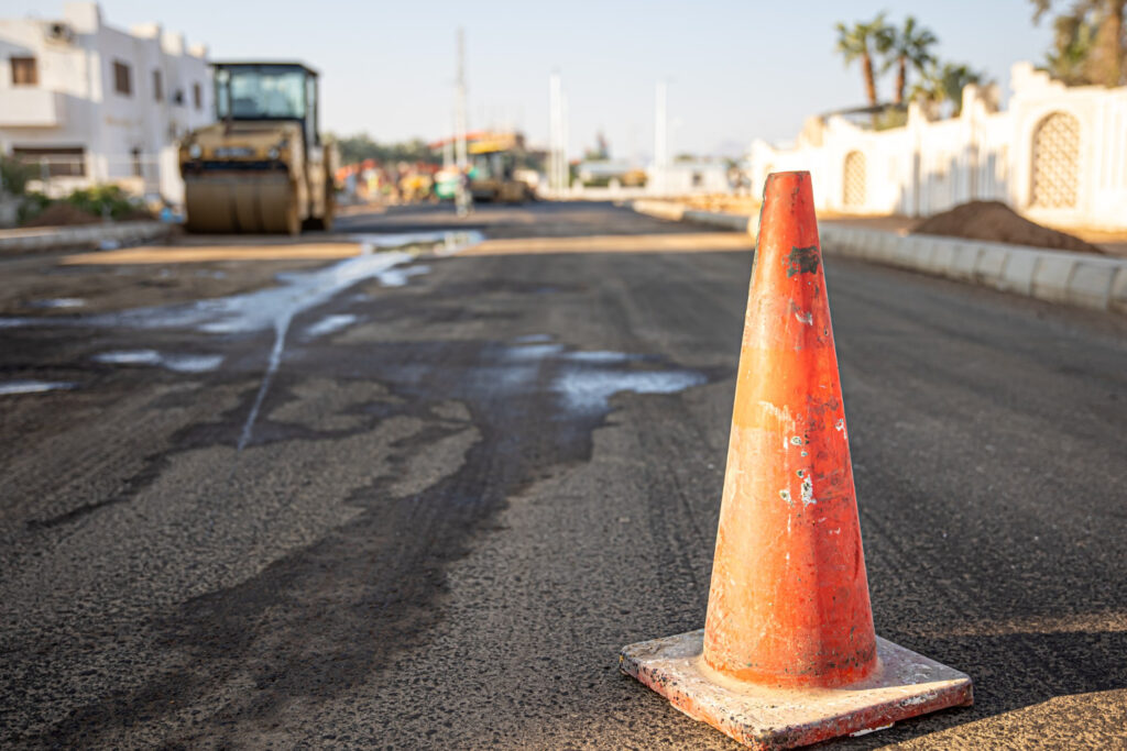 קונוס בטיחות כתום מסמן אזור עבודה בכביש בעיר, עם ציוד כבד ברקע.